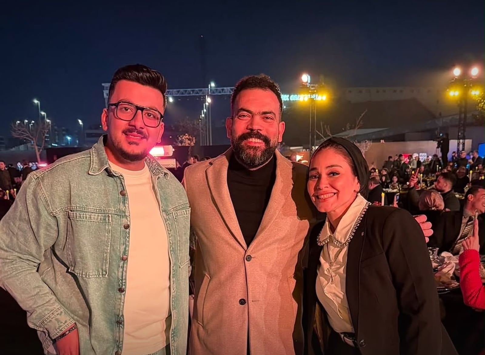Mohamed Amr Moharez with two guests at an evening event, floodlights and seated tables behind them.